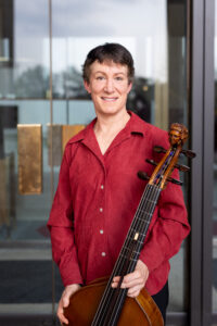 A woman in a red shirt stands in front of a glass door, holding a bass viol.