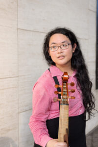 A woman with long dark hair wearing a pink shirt stands by a stone wall, holding a bass viol.