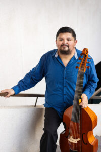 A man with dark hair and a beard stands along a wall, holding a bass viol.